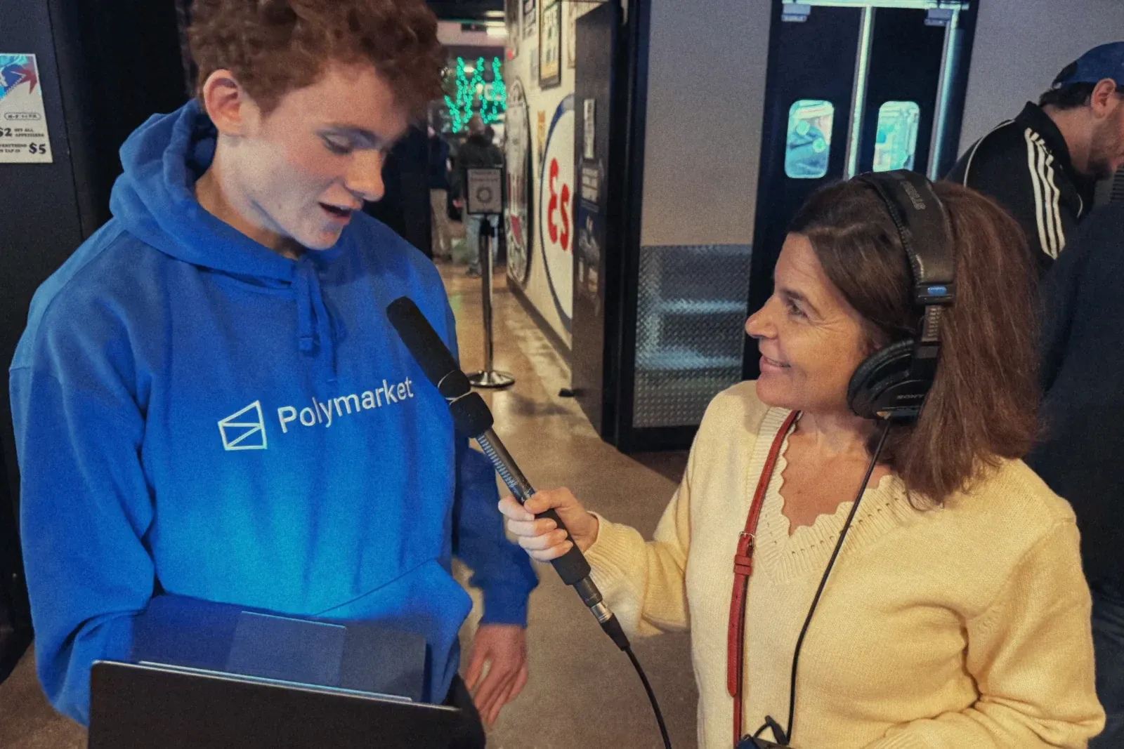 Ryan, in a Polymarket hoodie and holding a laptop, answers a radio reporter holding a microphone.
